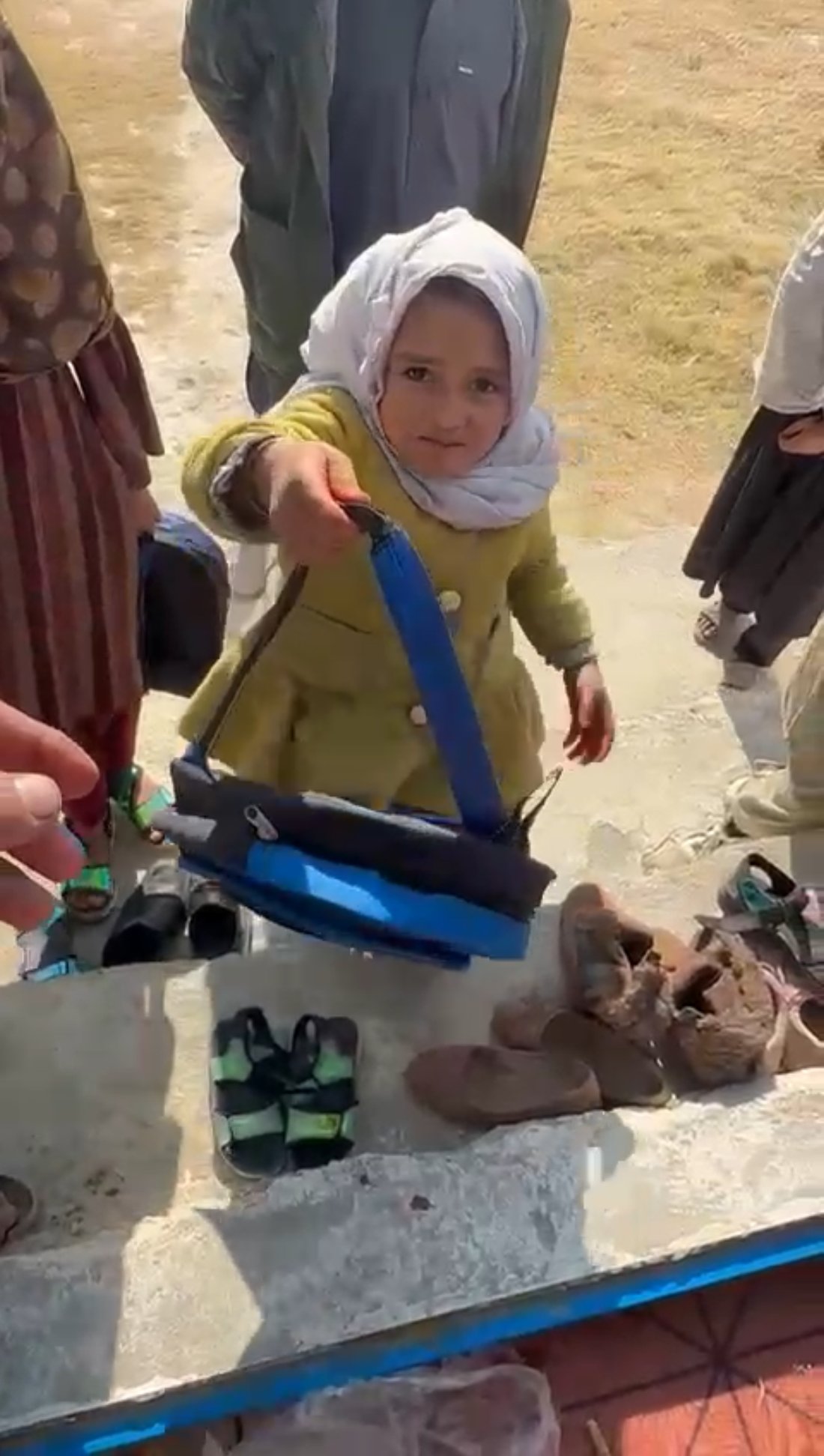 A young girl in a yellow coat and white headscarf holds up the blue backpack she has just received