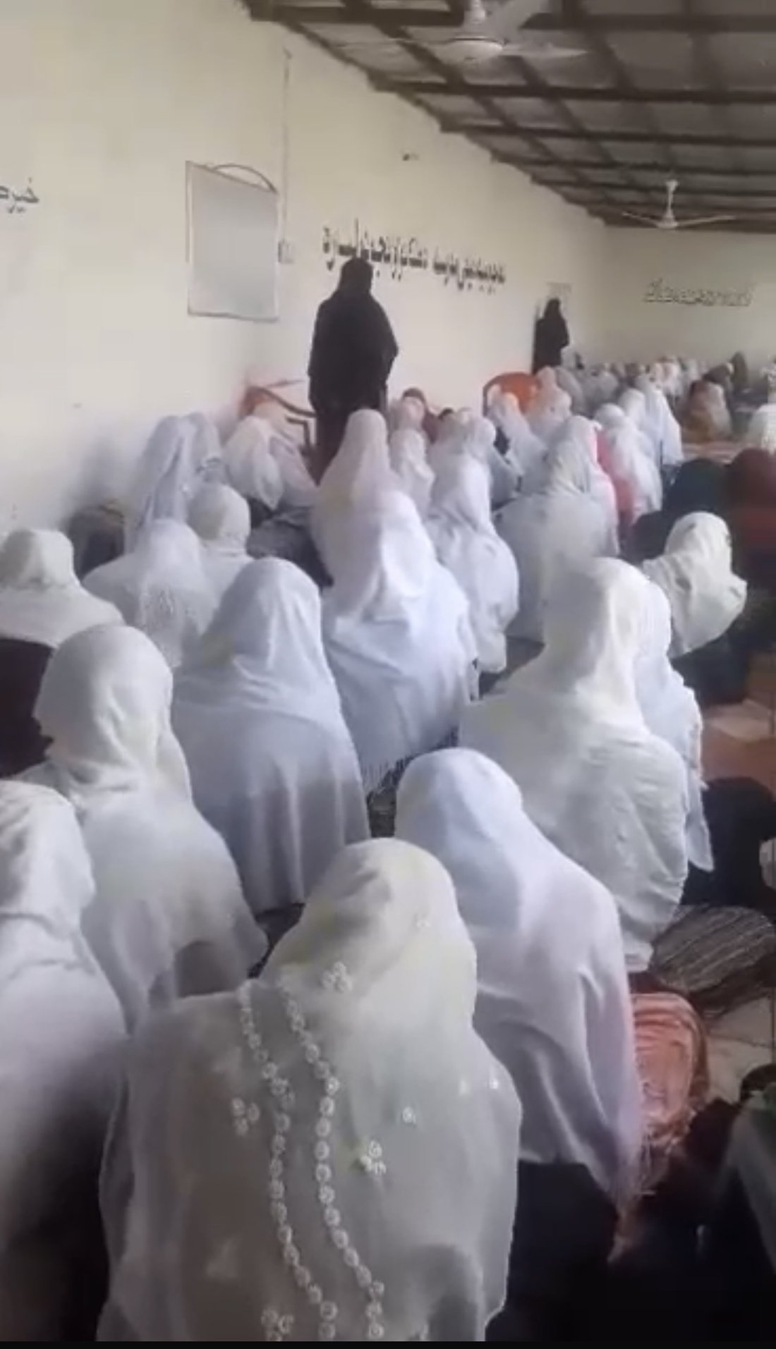 A full classroom of girls in white headscarves seated on the floor during a lesson