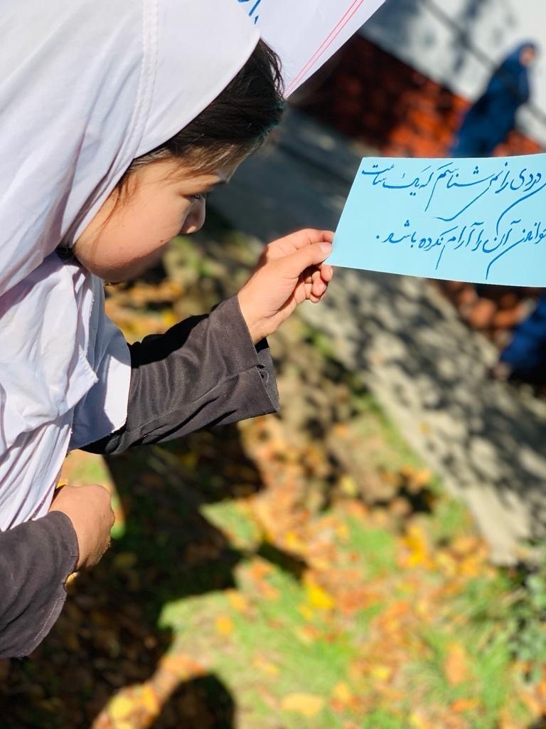 A girl in a white headscarf reads a handwritten note on blue paper, outdoors in autumn sunlight