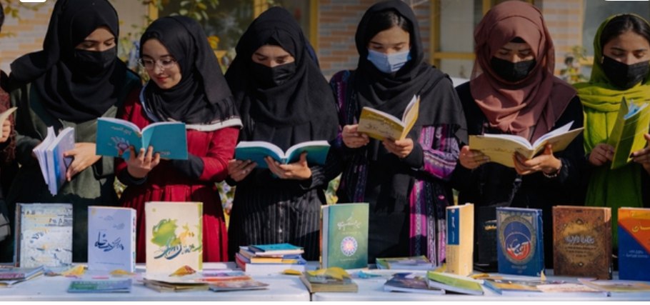 A row of students standing together reading open books, with more books laid out on a table in front of them