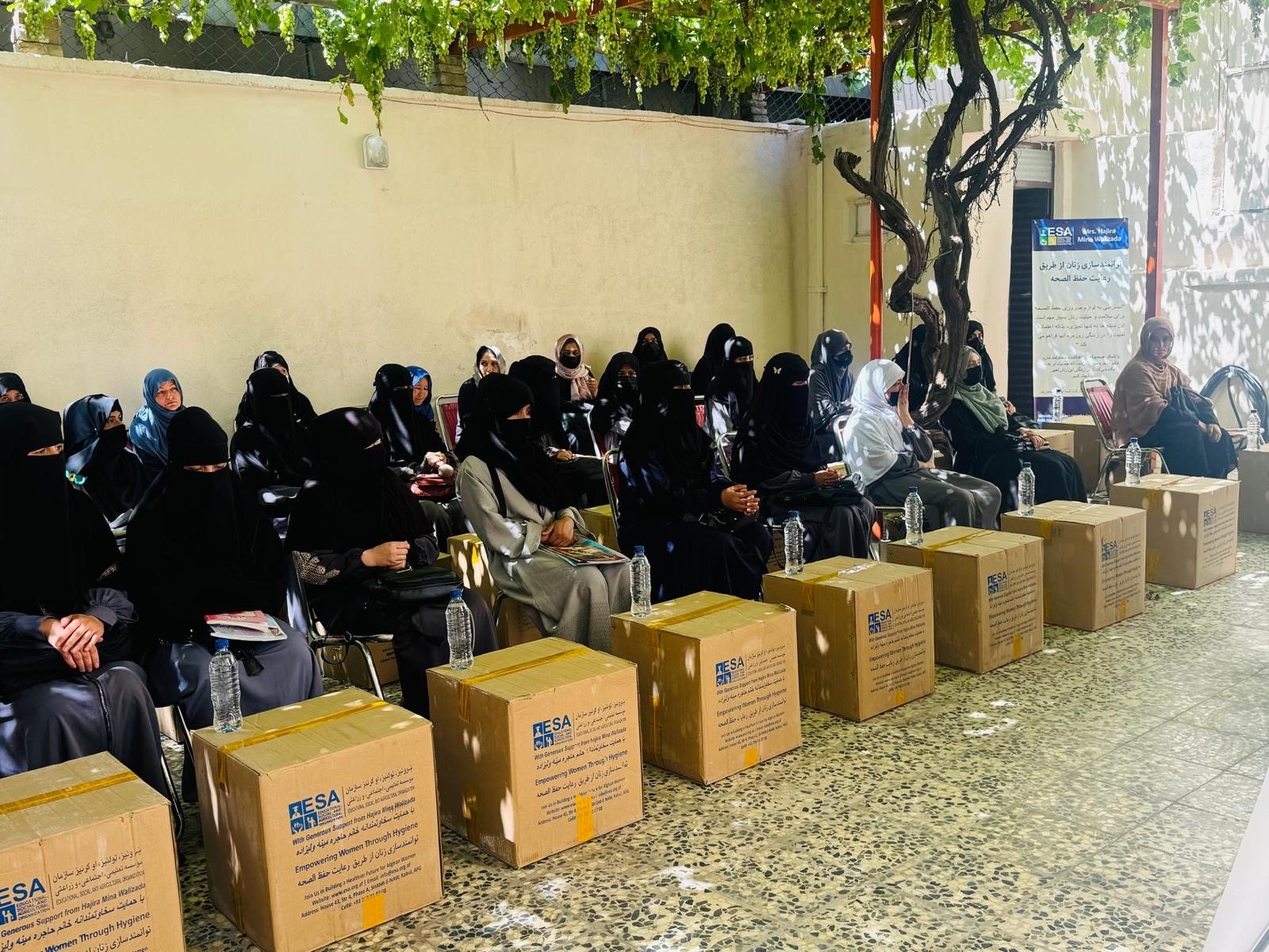 Women seated in rows at ESA's Kabul facility, each with a labelled ESA hygiene kit box beside her