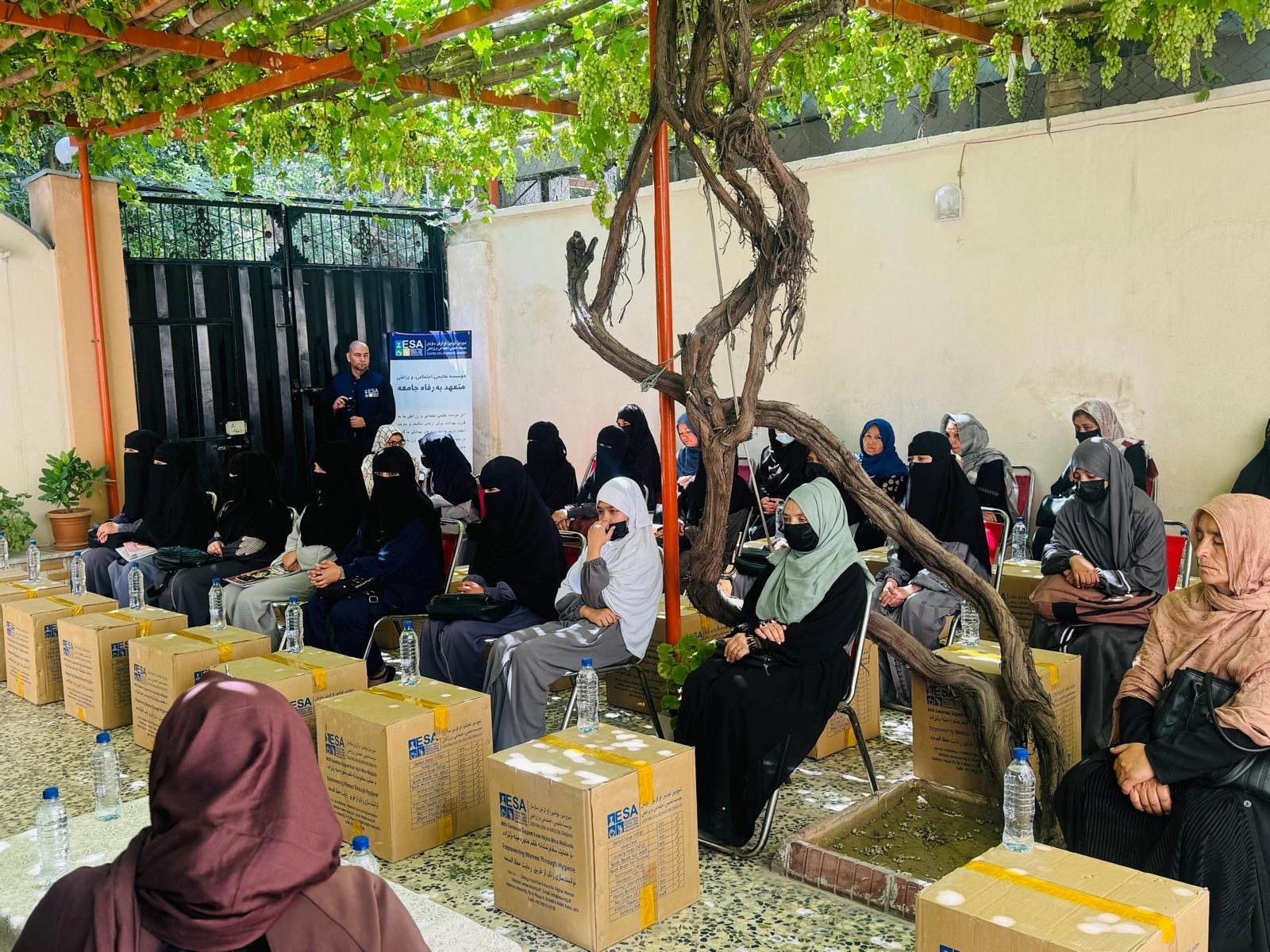 Women gathered under a grape arbour in ESA's Kabul courtyard, each seated beside a hygiene kit box, with ESA staff coordinating