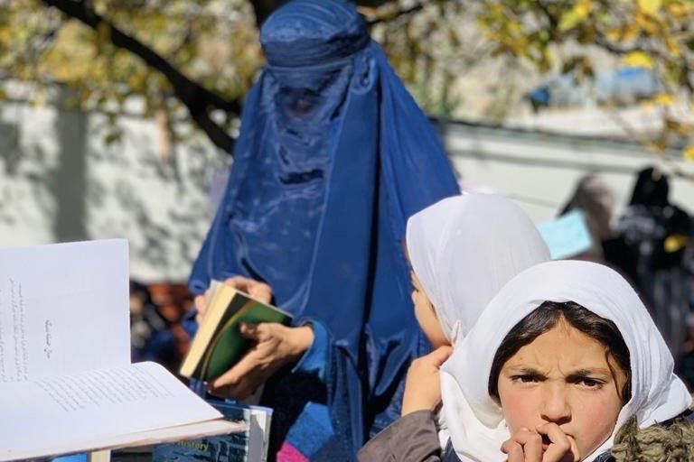 Girls seated outdoors with open books at a reading circle, one holding a book open in her hands